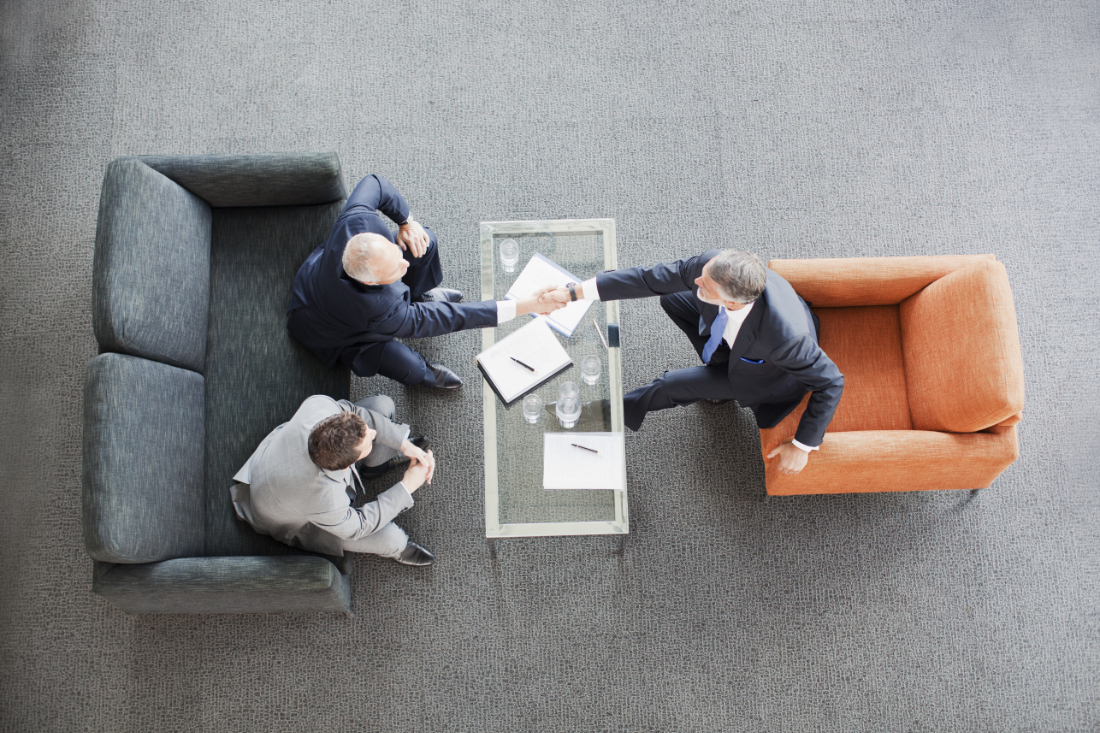Top view of three business professionals having a meeting, with two shaking hands across a glass table in a modern office setting.