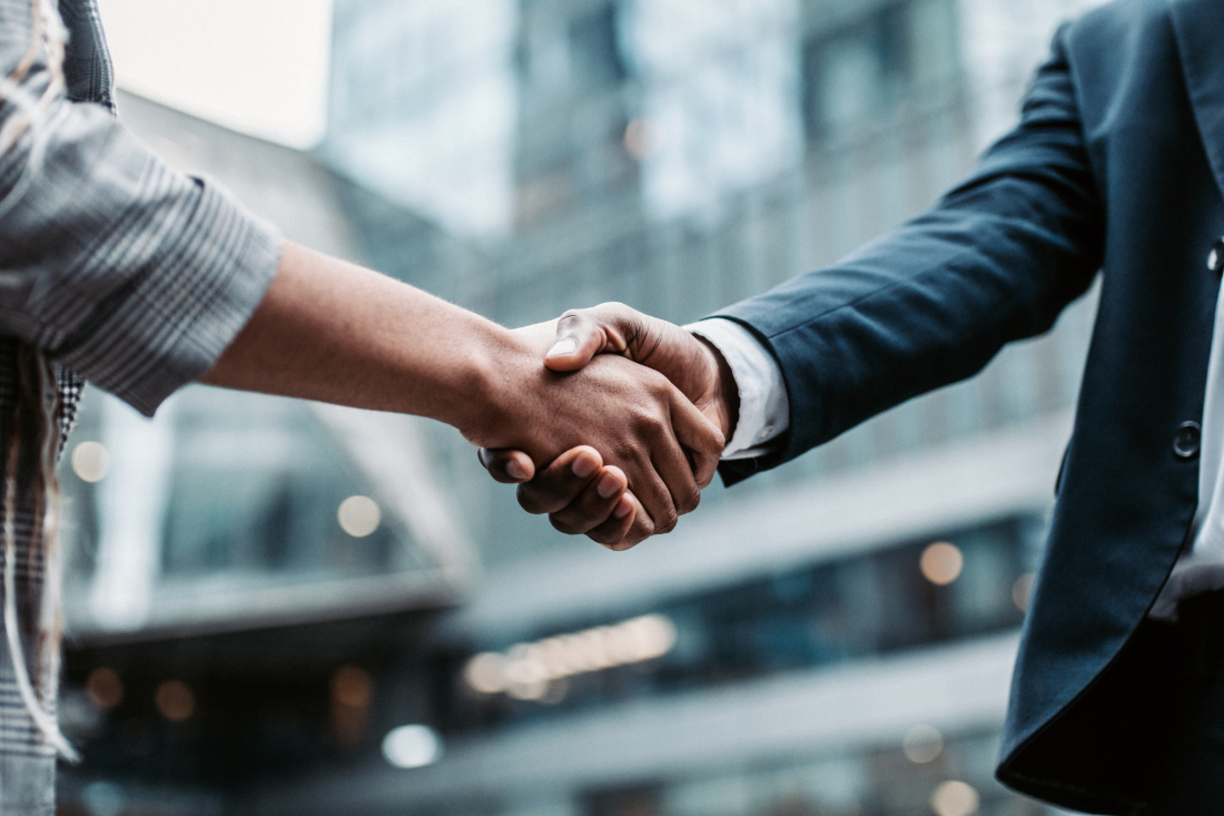 Close-up of a handshake between two business professionals, one in casual attire and the other in a formal suit, with modern office buildings in the background.