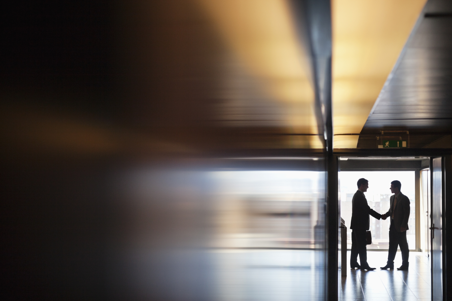 Two business professionals shaking hands in a modern office hallway with large windows and cityscape view in the background.